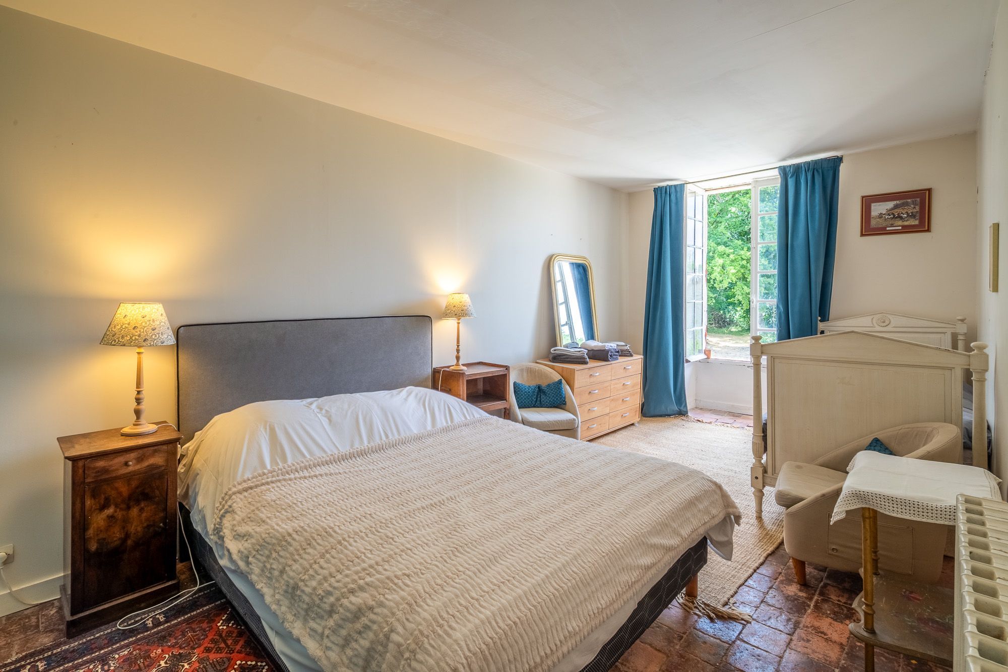 Ein Schlafzimmer mit einem Bett, Nachttischlampen, einer Kommode, einem Sessel, einem Spiegel und einem fenster mit Vorhängen.