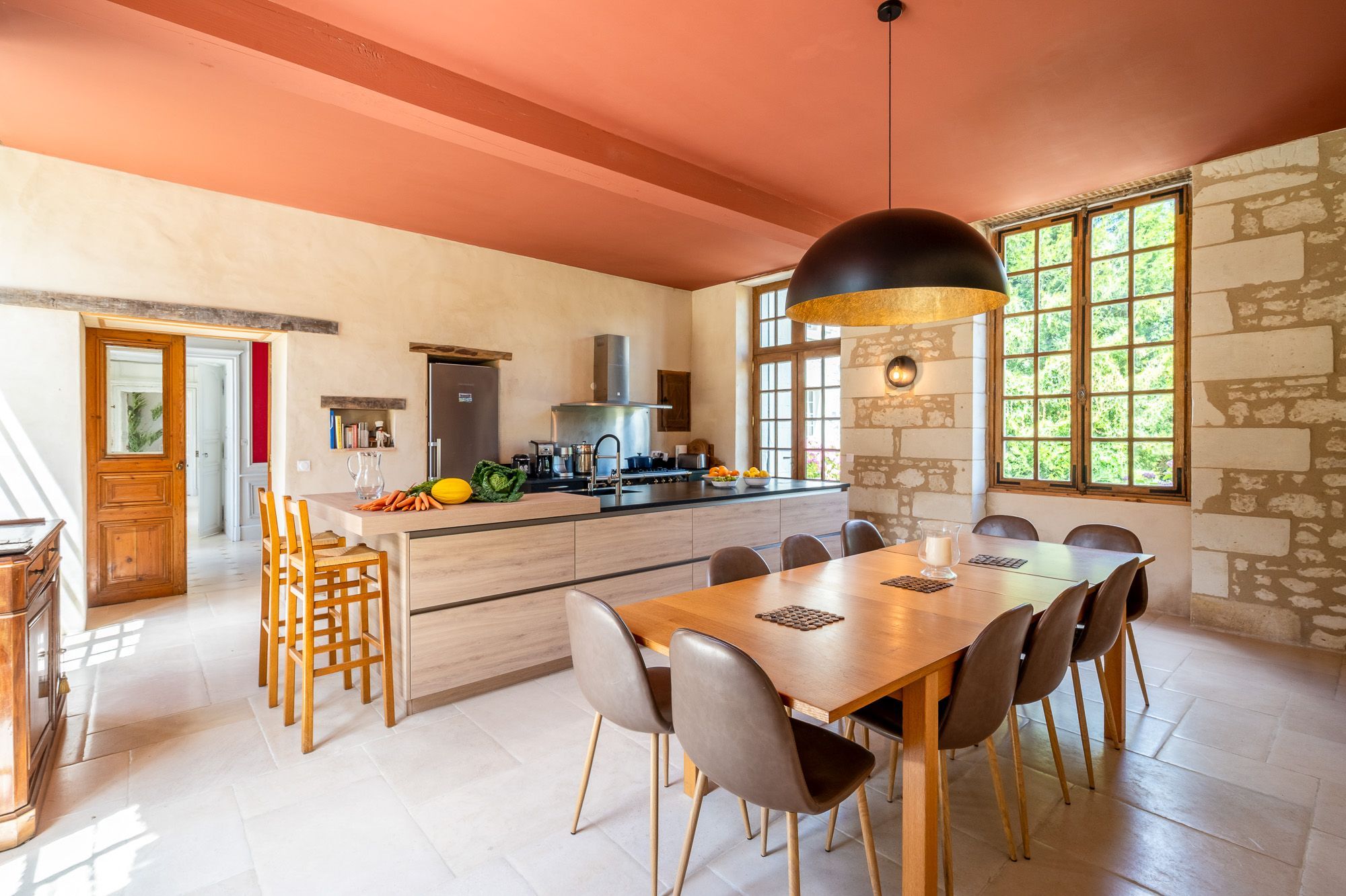 Een ruime keuken met een centraal eiland, een eethoek met een lange tafel en uitzicht op groen door grote ramen.