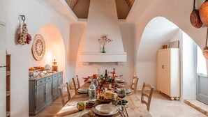 Comedor con mesa de madera dispuesta para una comida, zona de cocina con armarios e iluminación colgante.