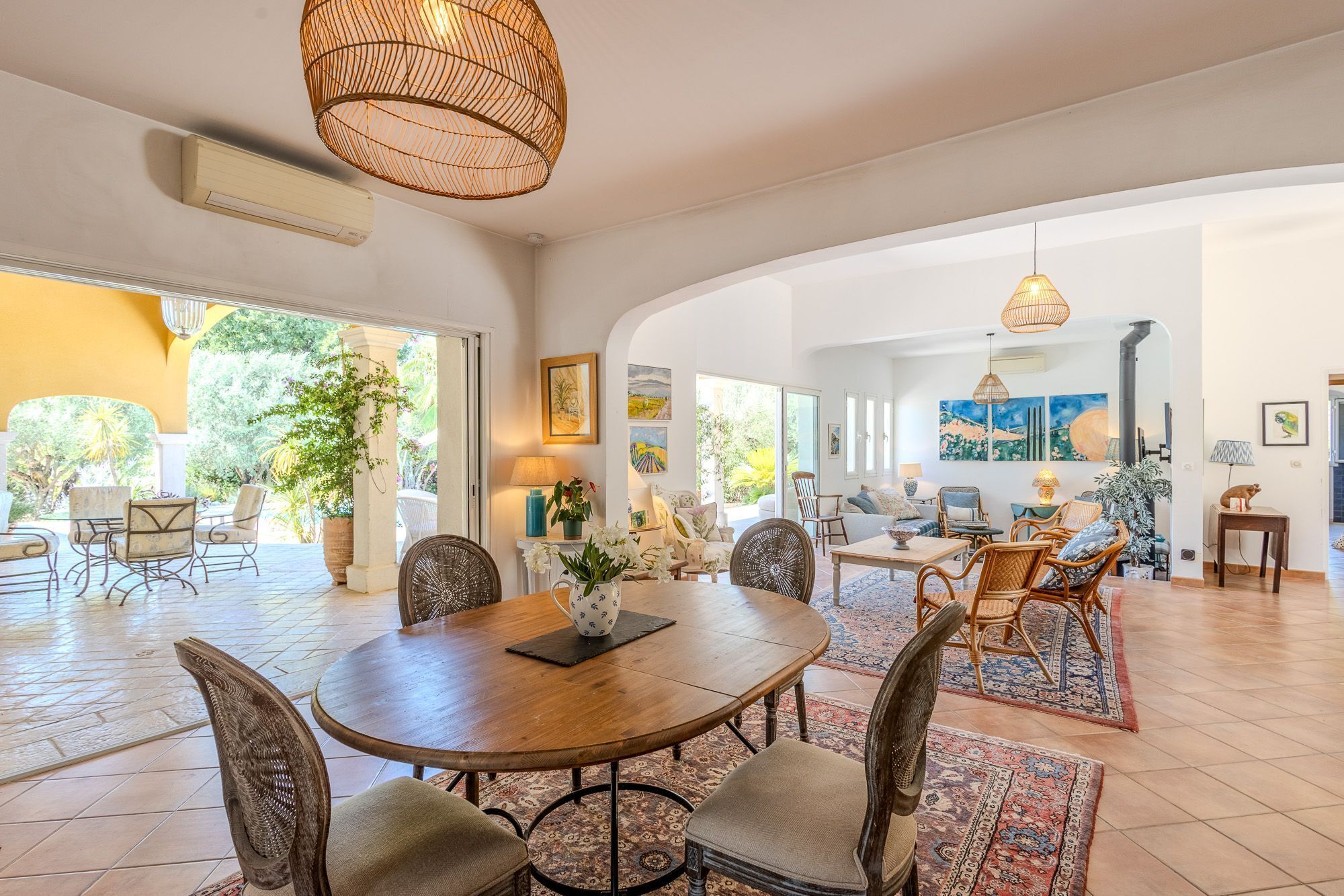 A spacious dining area with a round wooden table and wicker chairs, surrounded by large windows and doors leading to an outdoor patio.