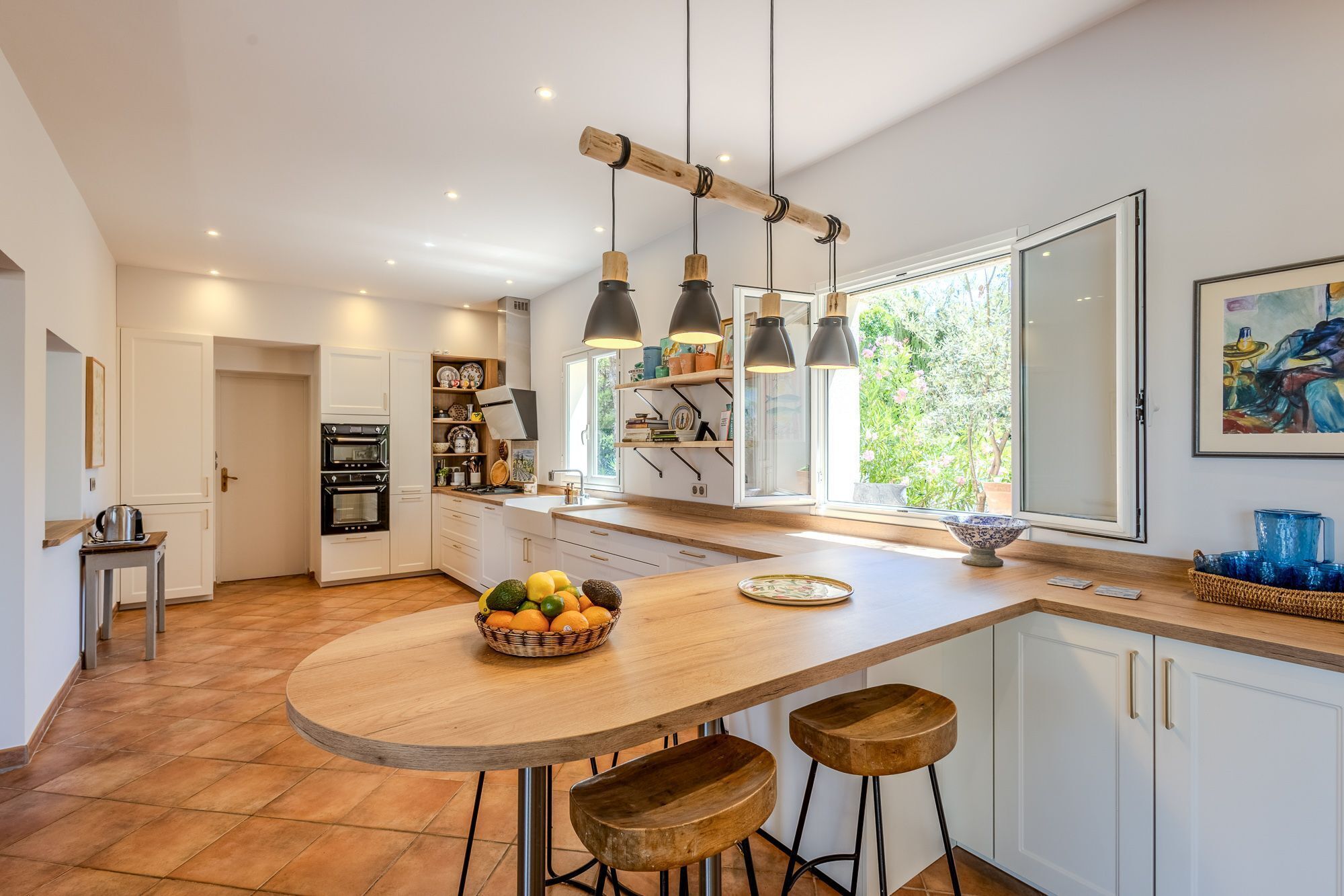A modern kitchen with a central island, pendant lighting, and a view of greenery outside.