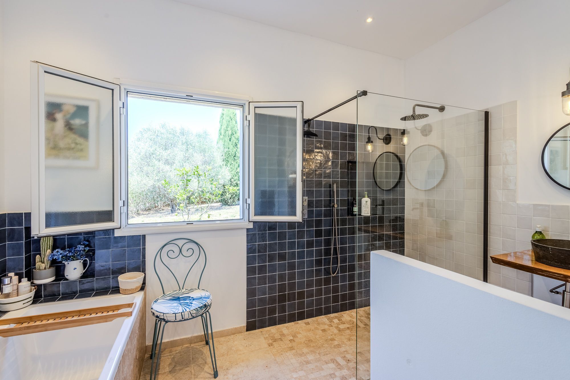 A bathroom with a shower, a window with a view of trees, a chair, and a sink.
