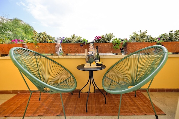 Two green wire mesh chairs, a small round table, a bottle, and a bowl of fruit on a balcony with yellow walls and potted plants.