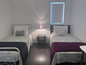 Ein kleiner, schummrig beleuchteter Raum mit zwei Betten, einem Nachttisch mit Lampe und einem Fenster mit blauem Licht.
