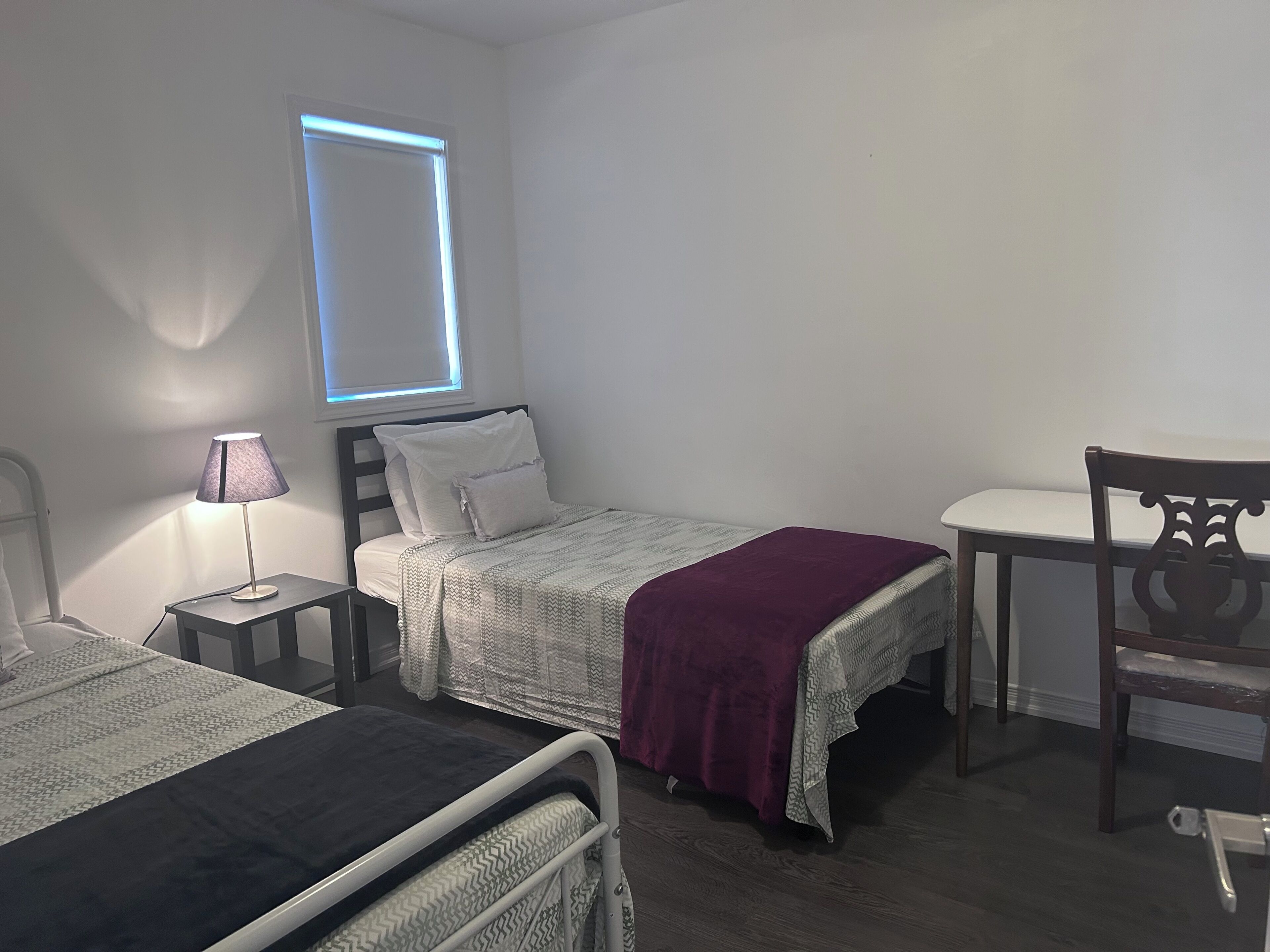 A bedroom with a bed, bedside table, lamp, chair, and a window with blinds.