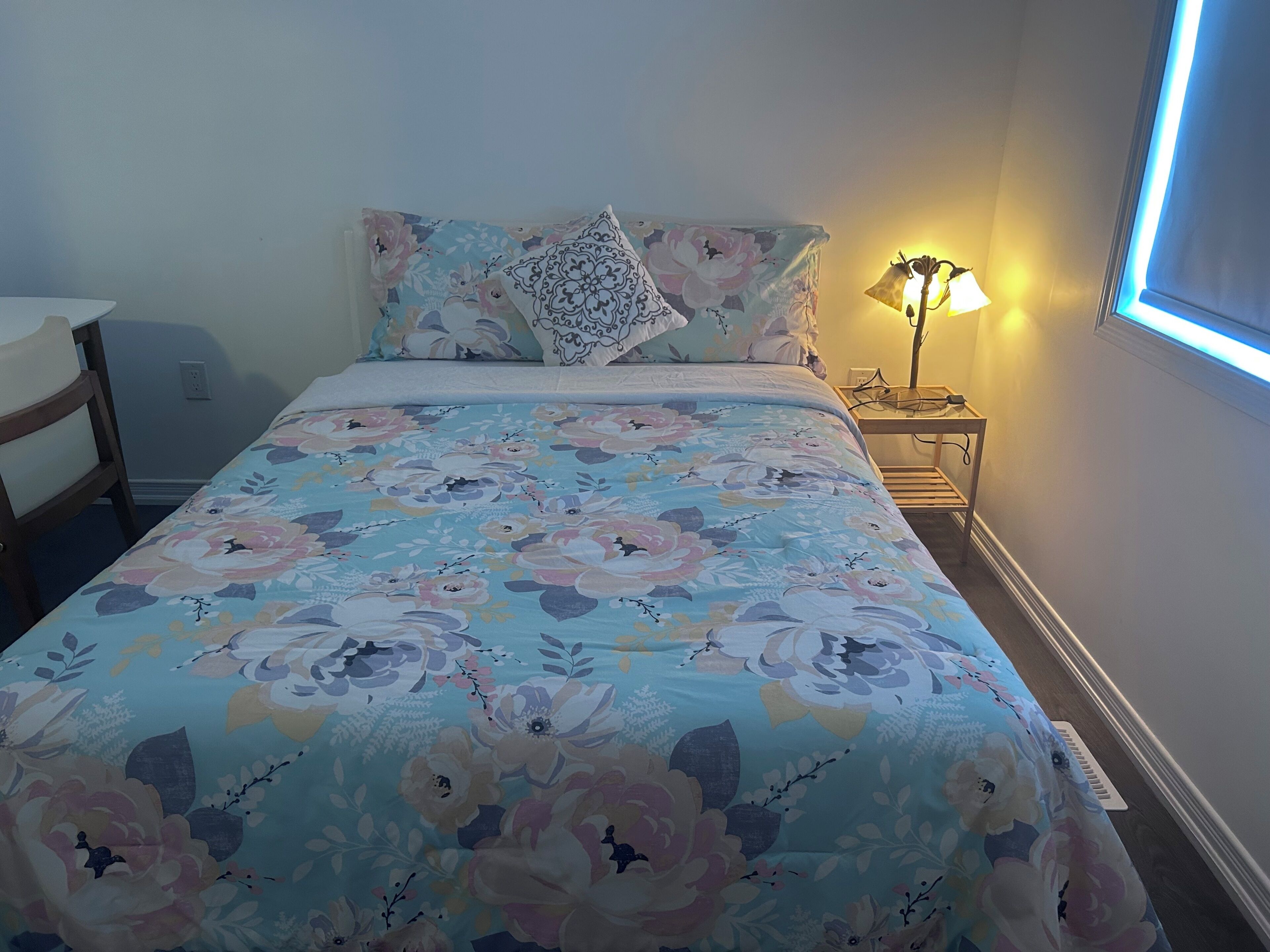 Une chambre à coucher avec une couvre-lit à motifs floraux, une lampe de chevet et une fenêtre avec des stores.