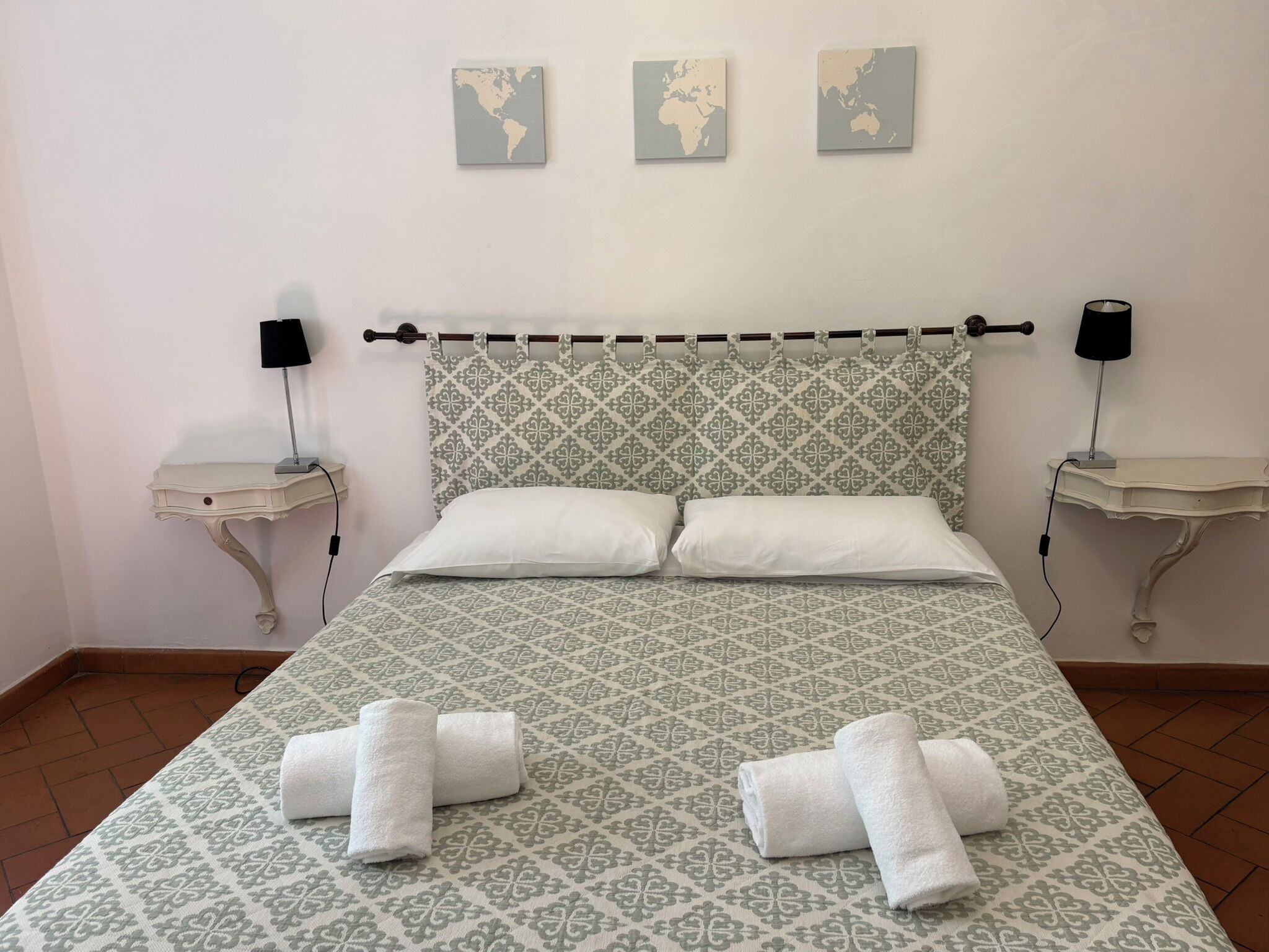 A neatly made bed with patterned bedding, two bedside tables with lamps, and three world maps on the wall.