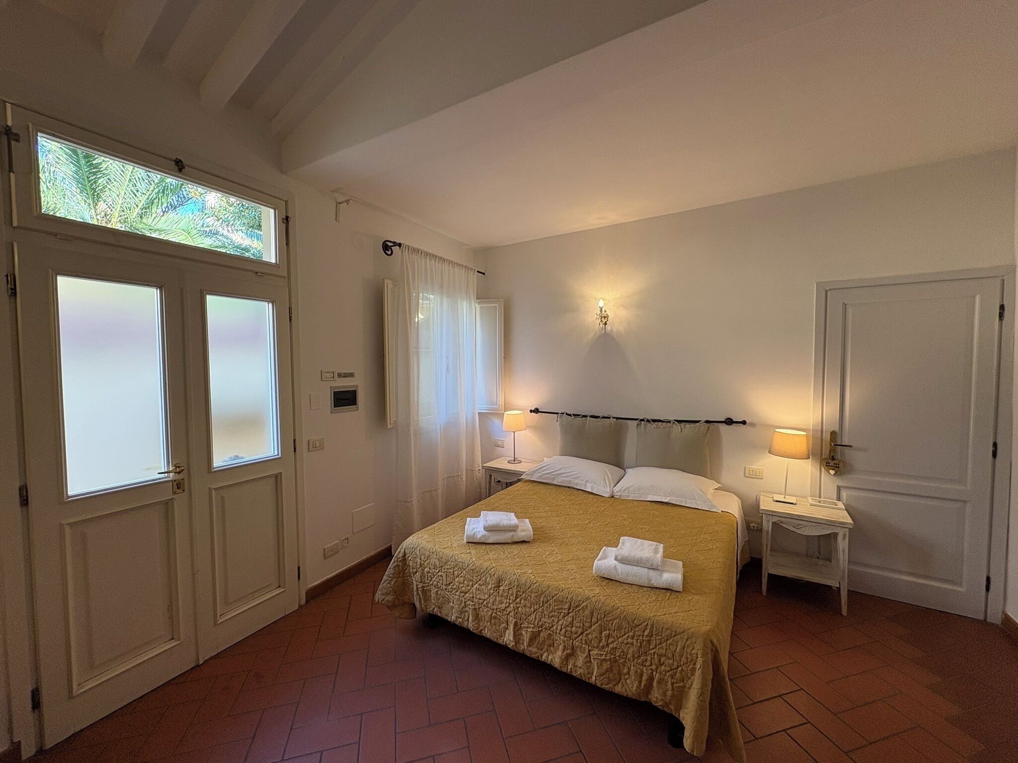 A hotel room with a bed, two bedside tables, a nightstand, a window with frosted glass, and a door.