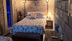 A bedroom with a brick wall, a bed with floral bedding, a ceiling fan, and a nightstand with a lamp. - La Casona de Los Angeles de San Juan (Taxco)