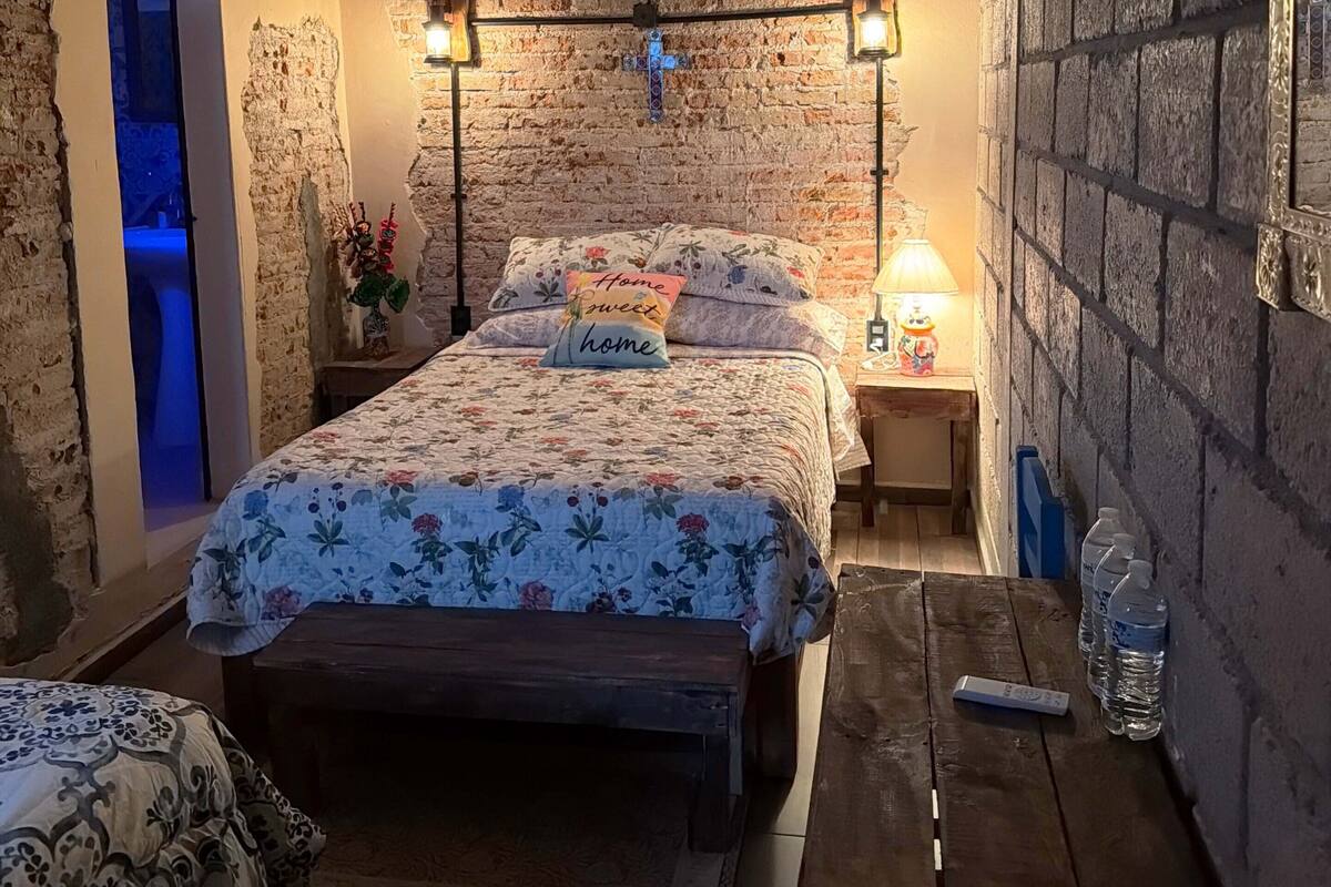 Ein Schlafzimmer mit Backsteinwand, einem Bett mit floralem Bettzeug, einem Deckenventilator und einem Nachttisch mit Lampe.