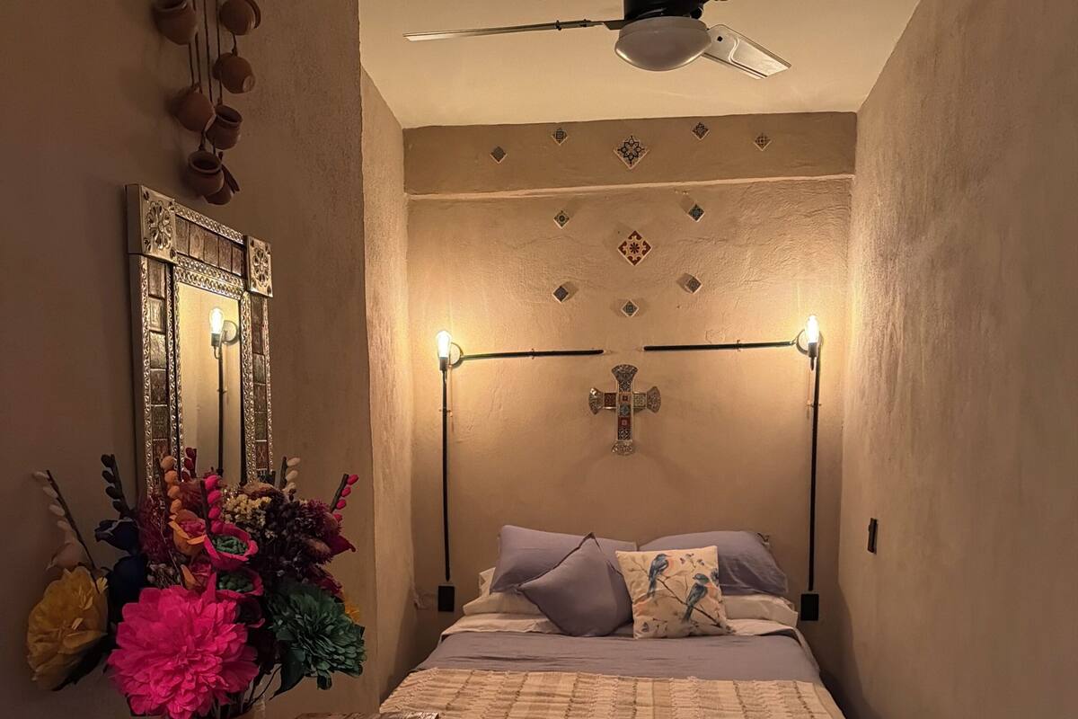 Ein Schlafzimmer mit einem Bett, einem Nachttisch mit Blumenvase, einem Deckenventilator und einem an der Wand befestigten Kreuz.