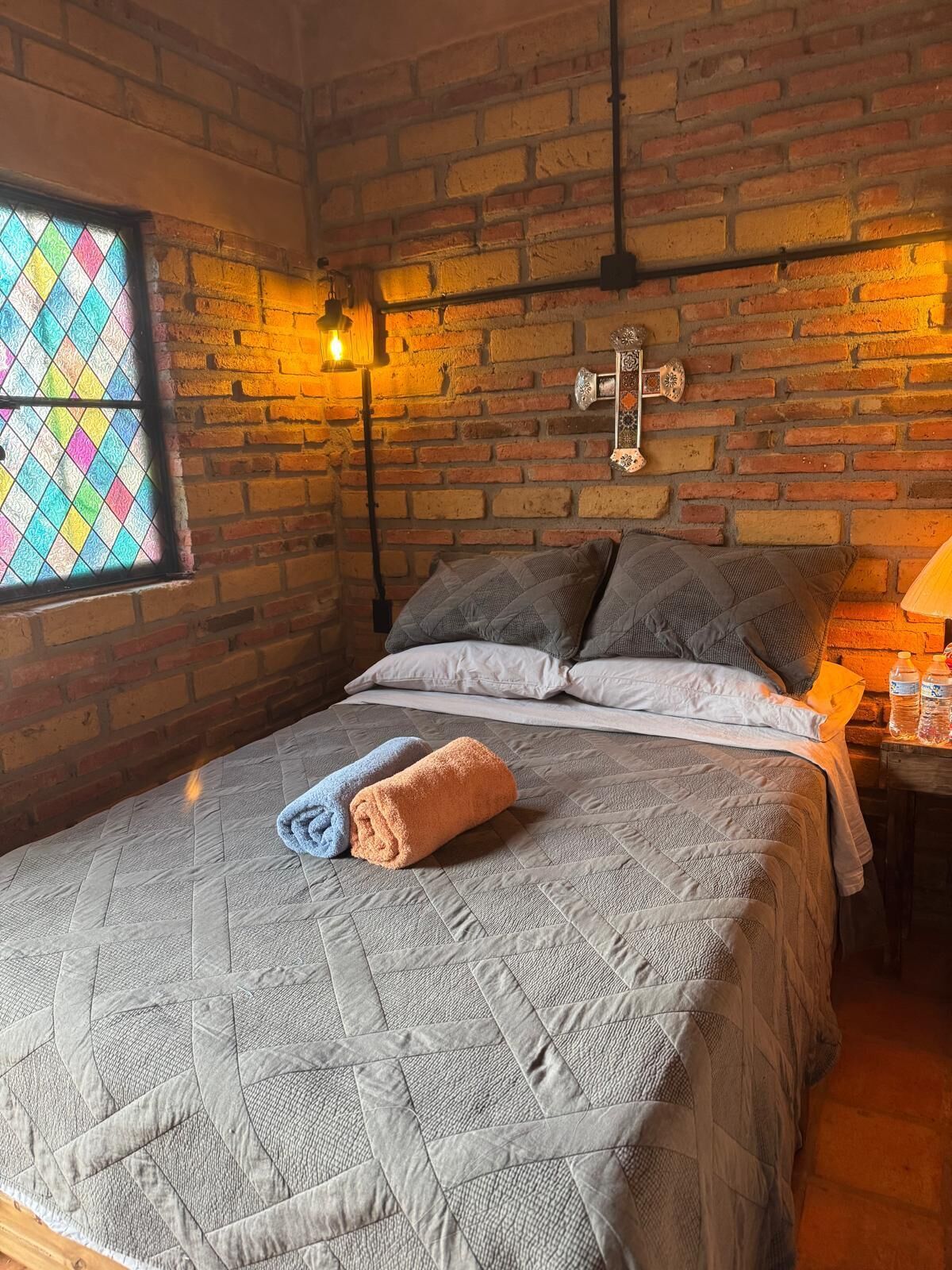 A room with a brick wall, a bed with a quilted bedspread, two pillows, a window with a stained glass design, and a wall-mounted light fixture.