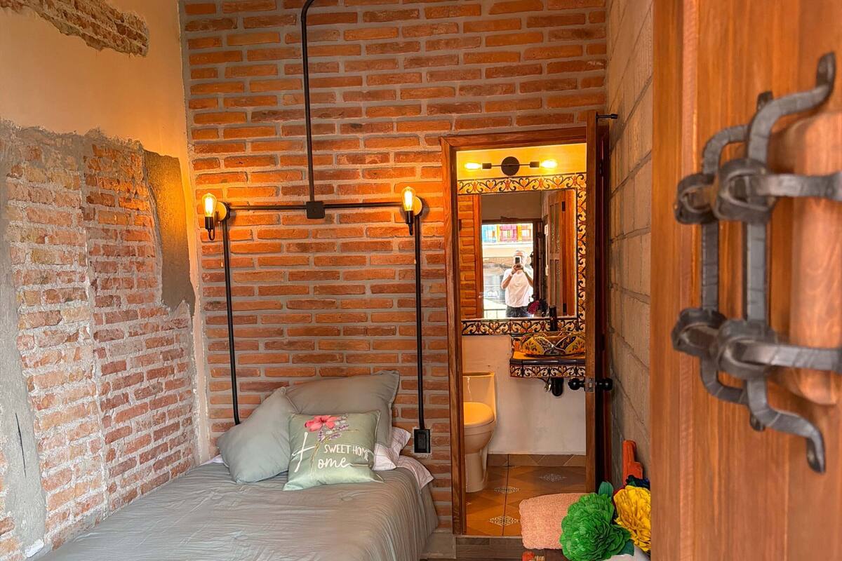 Ein Schlafzimmer mit Backsteinwand, Deckenventilator, Bett mit floralem Überwurf, Nachttisch mit Lampe und Holztür.