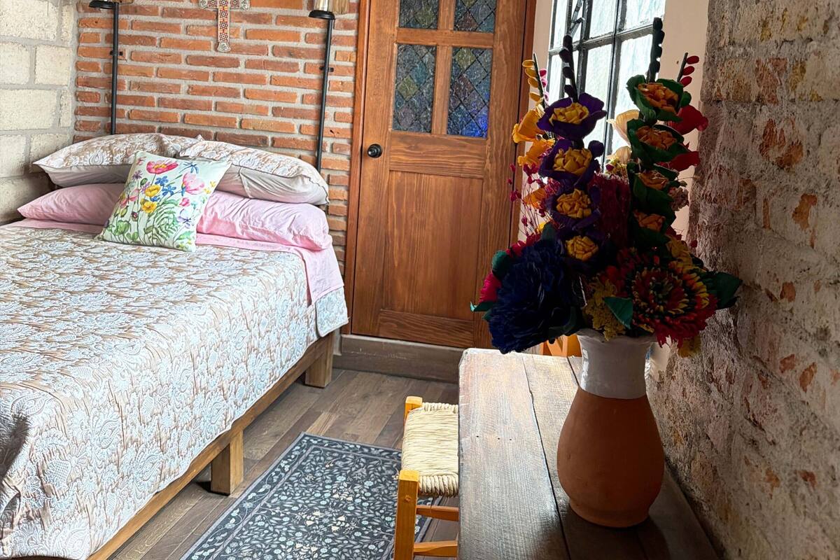 Ein Zimmer mit Backsteinwand, einer Holztür, einem Bett mit floralem Bettbezug, einem Holzstuhl, einer Vase mit Blumen und einem schwarz-weißen Teppich.