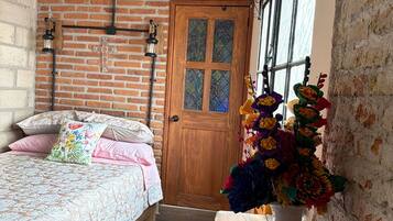 A room with a brick wall, a wooden door, a bed with floral bedding, a wooden chair, a vase with flowers, and a black and white rug.