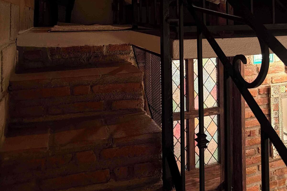 Eine Treppe mit schmiedeeisernem Geländer führt zu einem Schlafzimmer mit floraler Bettdecke und einem Nachttisch mit Lampe.