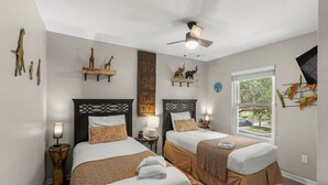 Ein Hotelzimmer mit zwei Betten, einem Deckenventilator, Kunstwerken an den Wänden, einem Fenster mit Blick und einem kleinen Tisch mit Lampe.