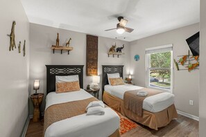 Ein Hotelzimmer mit zwei Betten, einem Deckenventilator, Kunstwerken an den Wänden, einem Fenster mit Blick und einem kleinen Tisch mit Lampe.