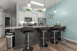 Eine moderne Küche mit weißen Schränken, einem Edelstahl-Kühlschrank, einer Mikrowelle und einer Kaffeemaschine. Es gibt drei schwarze Barhocker und einen Mülleimer.