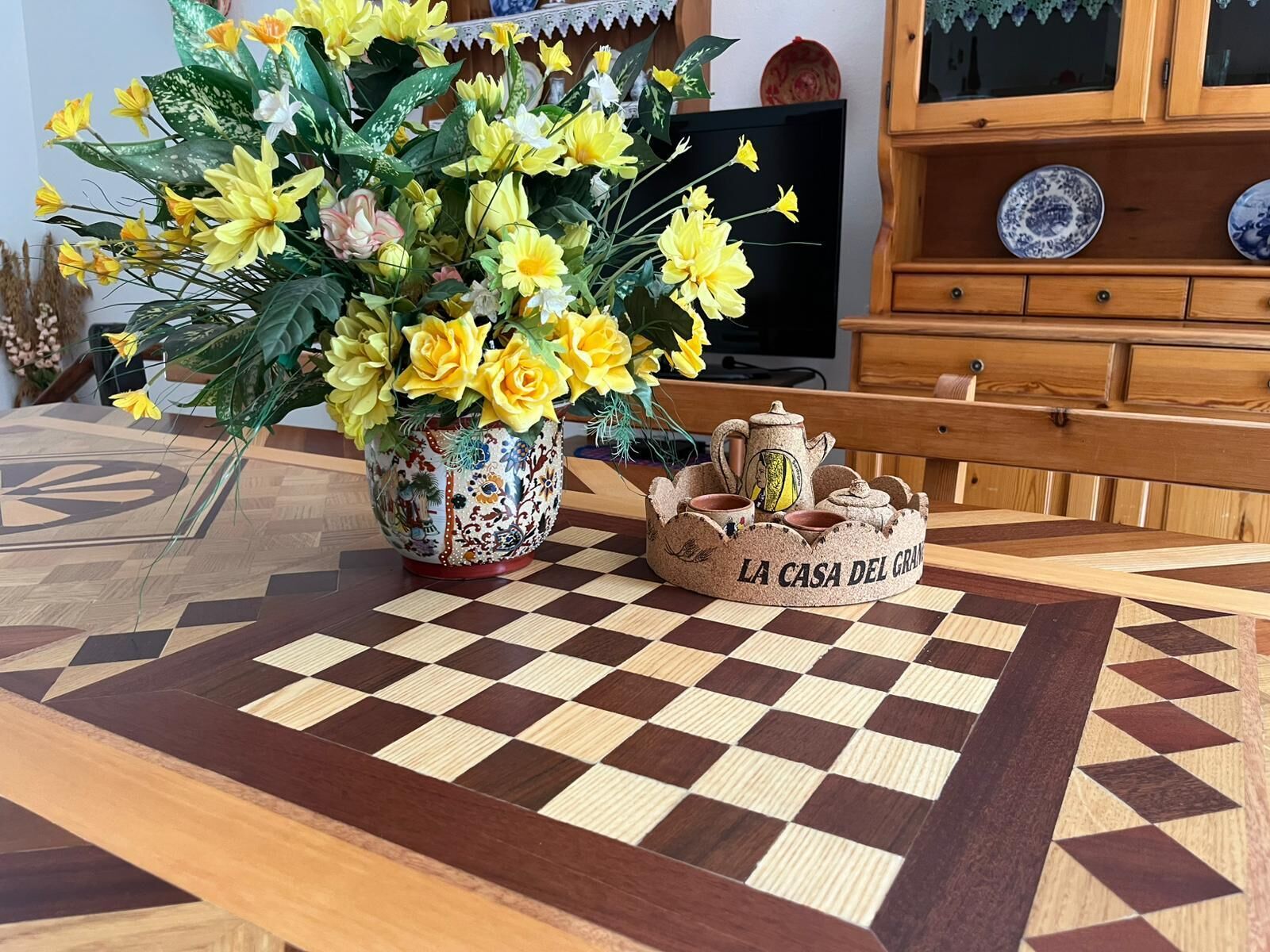 Un échiquier agrémenté d’un élément décoratif central, un buffet en bois contenant de la vaisselle, et un téléviseur en arrière-plan.