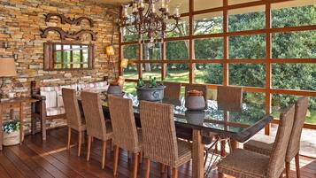 A dining area with a stone wall, wooden floor, wicker chairs, a glass table, and a view of greenery through large windows.
