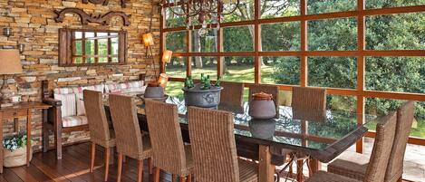 A dining area with a stone wall, wooden floor, wicker chairs, a glass table, and a view of greenery through large windows.