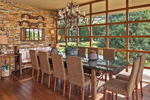 A dining area with a stone wall, wooden floor, wicker chairs, a glass table, and a view of greenery through large windows.
