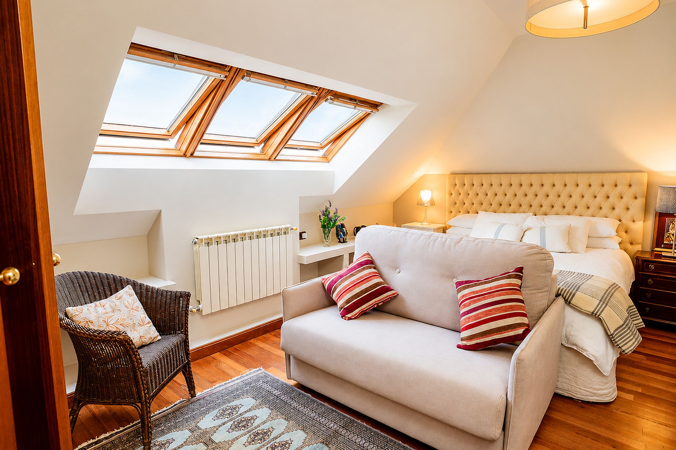 A cozy bedroom with a sloped ceiling, a bed with a tufted headboard, a sofa, a wicker chair, and a skylight.