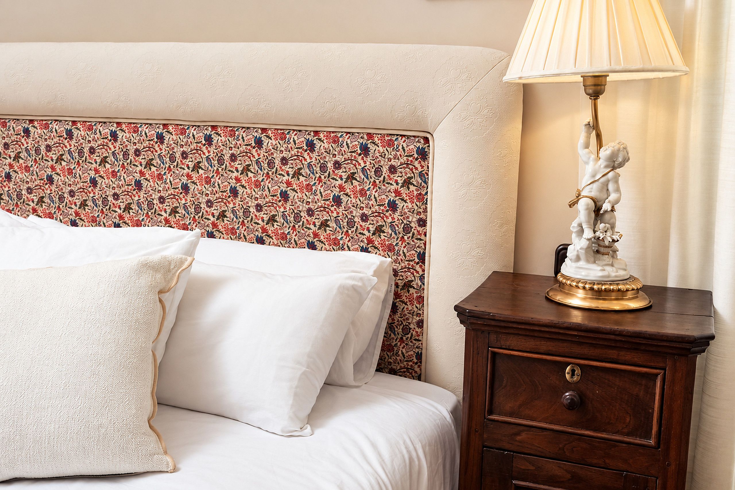 A bed with white linens, a floral-patterned headboard, and a nightstand with a lamp and a small statue.