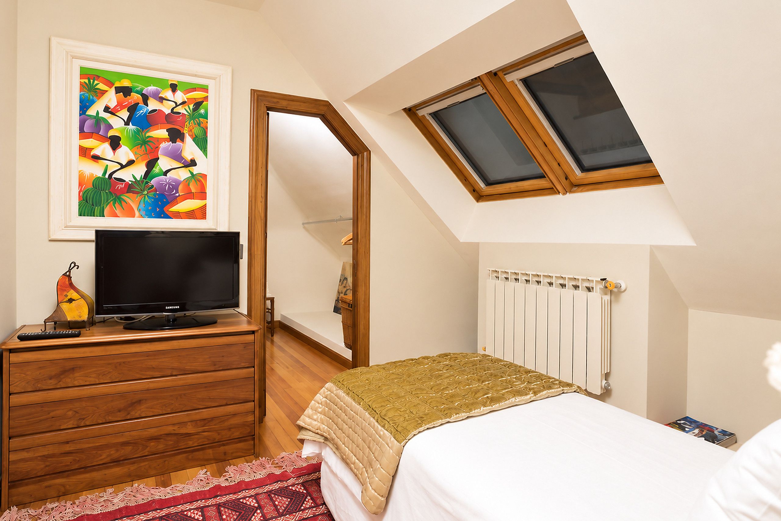 A bedroom with a bed, a television on a wooden dresser, a painting, a window, and a radiator.