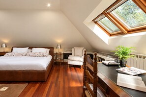 Une chambre avec un grand lit, un bureau avec une chaise, une petite table avec une lampe et une fenêtre donnant sur un paysage verdoyant.