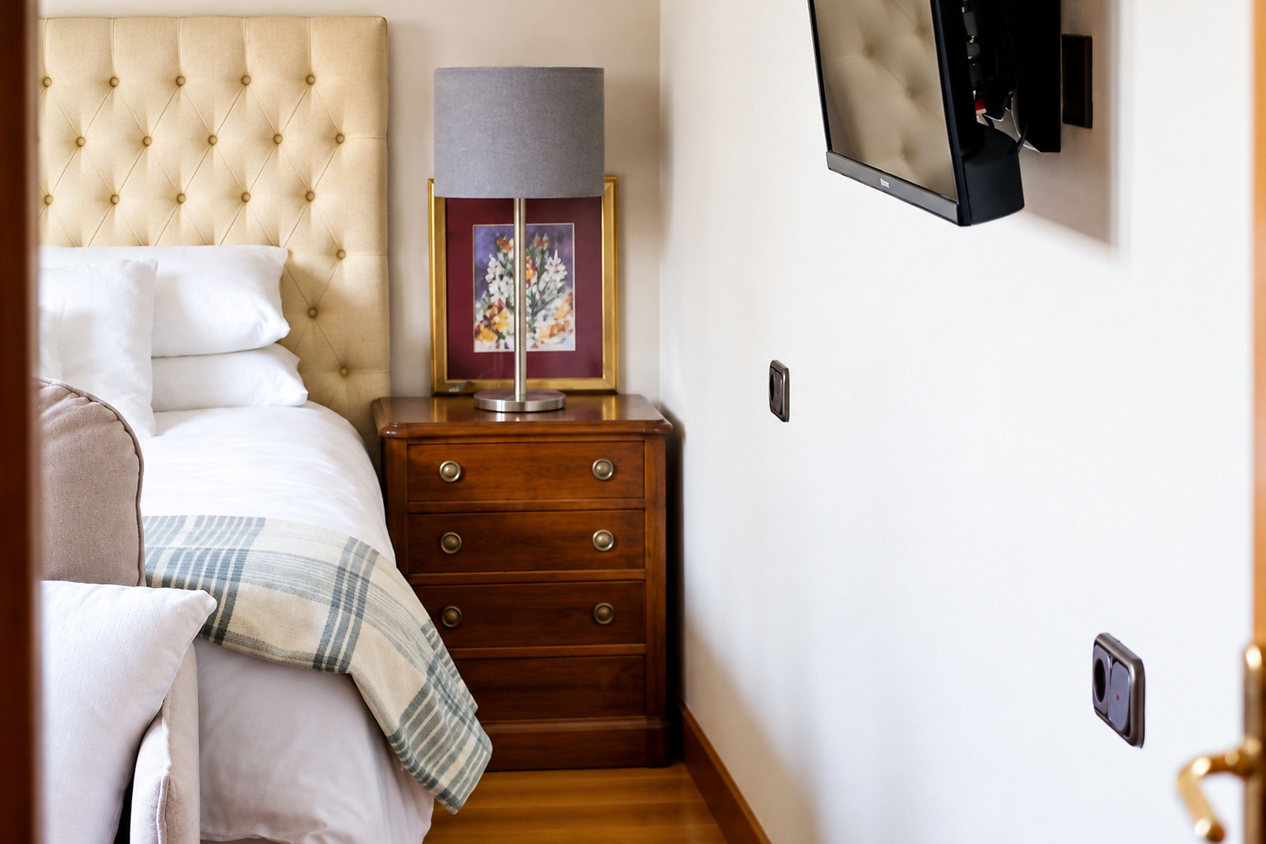 Quarto com cabeceira estofada, mesa de cabeceira com abajur e quadro emoldurado, cómoda de madeira e TV fixada na parede.