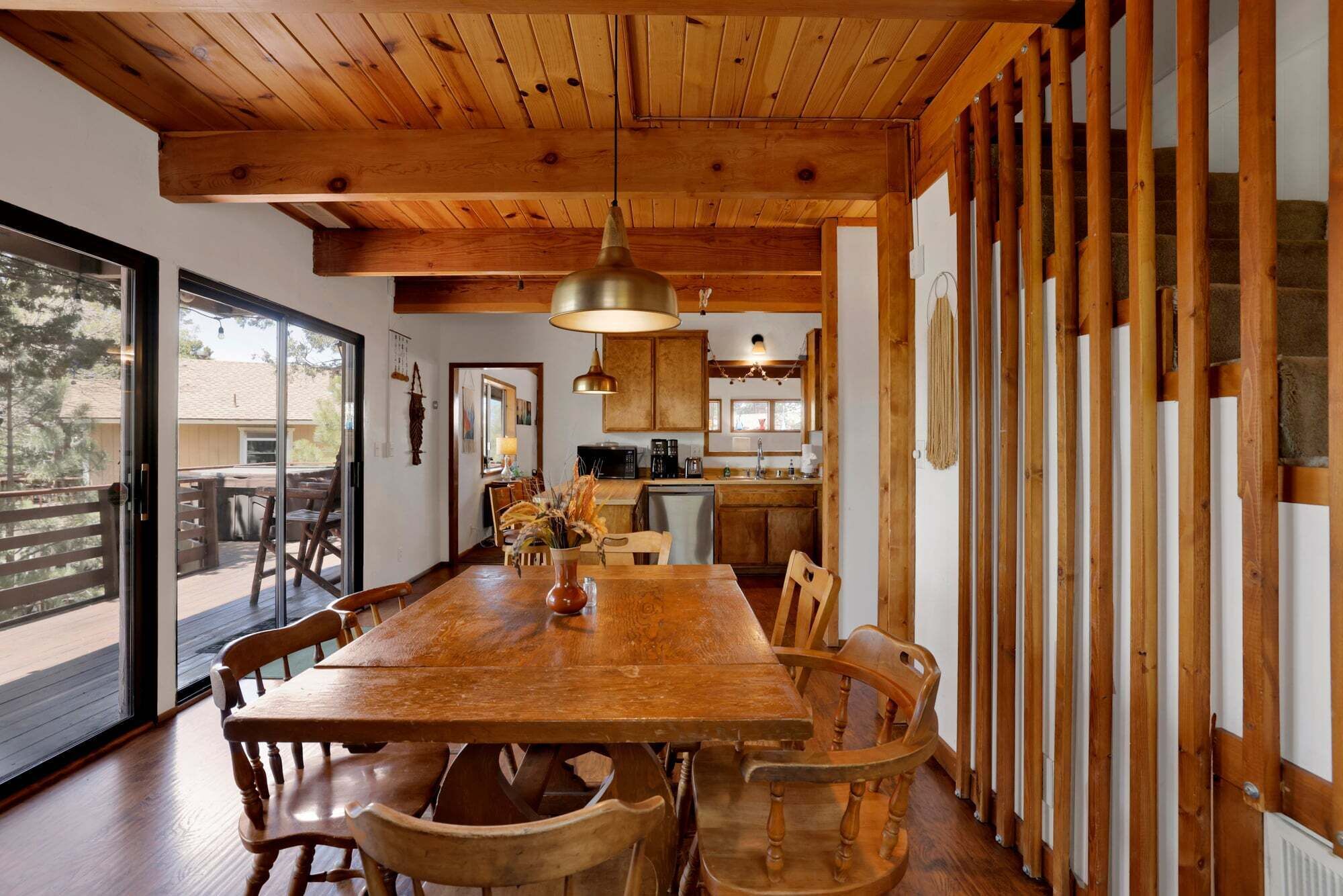 A wooden dining room with a large table and chairs, a hanging light fixture, and a view through a glass door to the outside.
