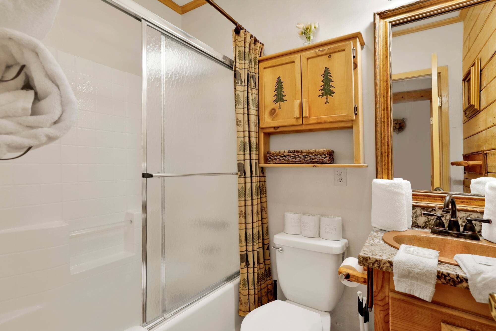 Un baño con cabina de ducha de vidrio, inodoro, lavabo con espejo y armarios de madera con diseños de árboles.