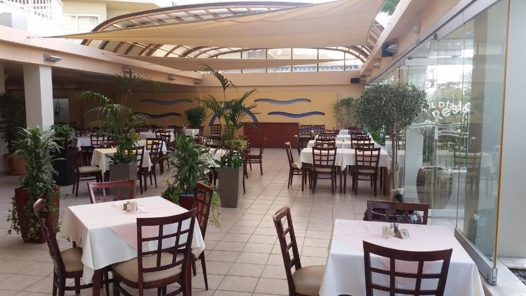 A spacious indoor dining area with multiple tables and chairs, potted plants, and a glass wall offering a view of the outside.