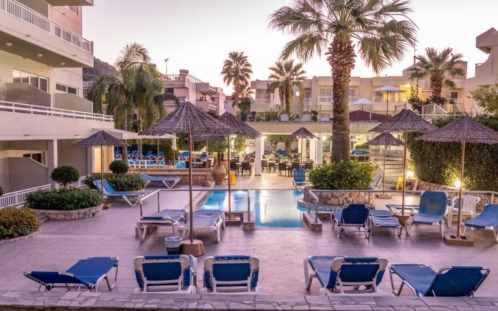 Ett poolområde med solstolar och parasoller, omgivet av palmer och byggnader.