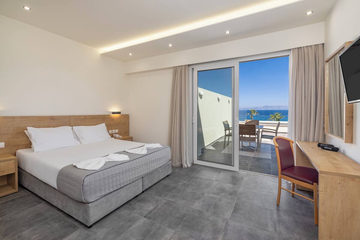 A hotel room with a bed, a desk, a chair, a TV, and a balcony with a view of the sea.