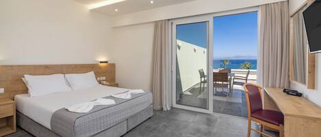 A hotel room with a bed, a desk, a chair, a TV, and a balcony with a view of the sea.