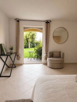Ein Schlafzimmer mit einem Bett, einem Sessel, einem Tisch und Blick auf einen Garten durch eine offene Tür.