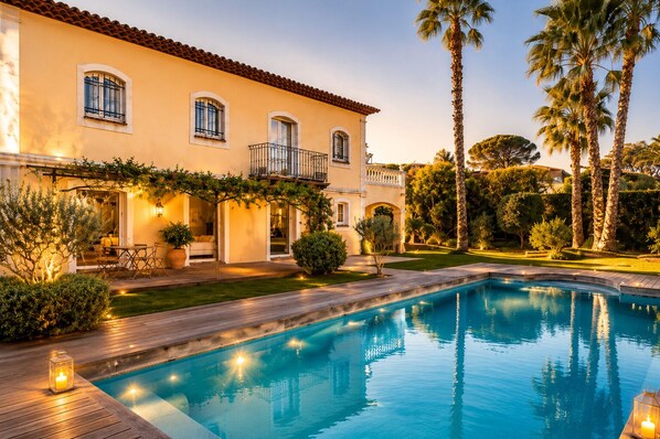 Vue sur une maison de style méditerranéen avec piscine, espace salon extérieur et un jardin luxuriant, le tout vu depuis le bord de la piscine.