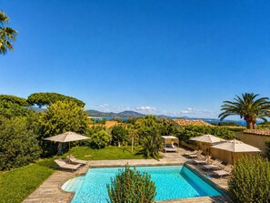 Ein Pool mit Liegestühlen und Sonnenschirmen, umgeben von Grün und Blick aufs Meer.