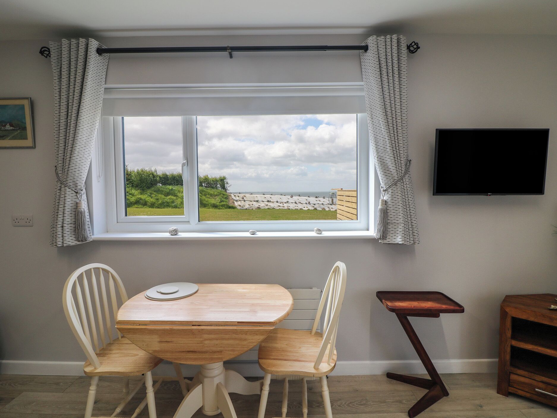 A room with a wooden table and chairs, a window with curtains, a flat-screen TV, and a small wooden side table.