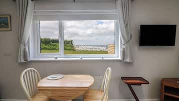 A room with a wooden table and chairs, a window with curtains, a flat-screen TV, and a small wooden side table.