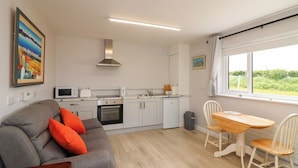 Een kleine keuken met een magnetron, broodrooster en oven, een grijze bank met oranje kussens, en een eethoek met een houten tafel en stoelen.