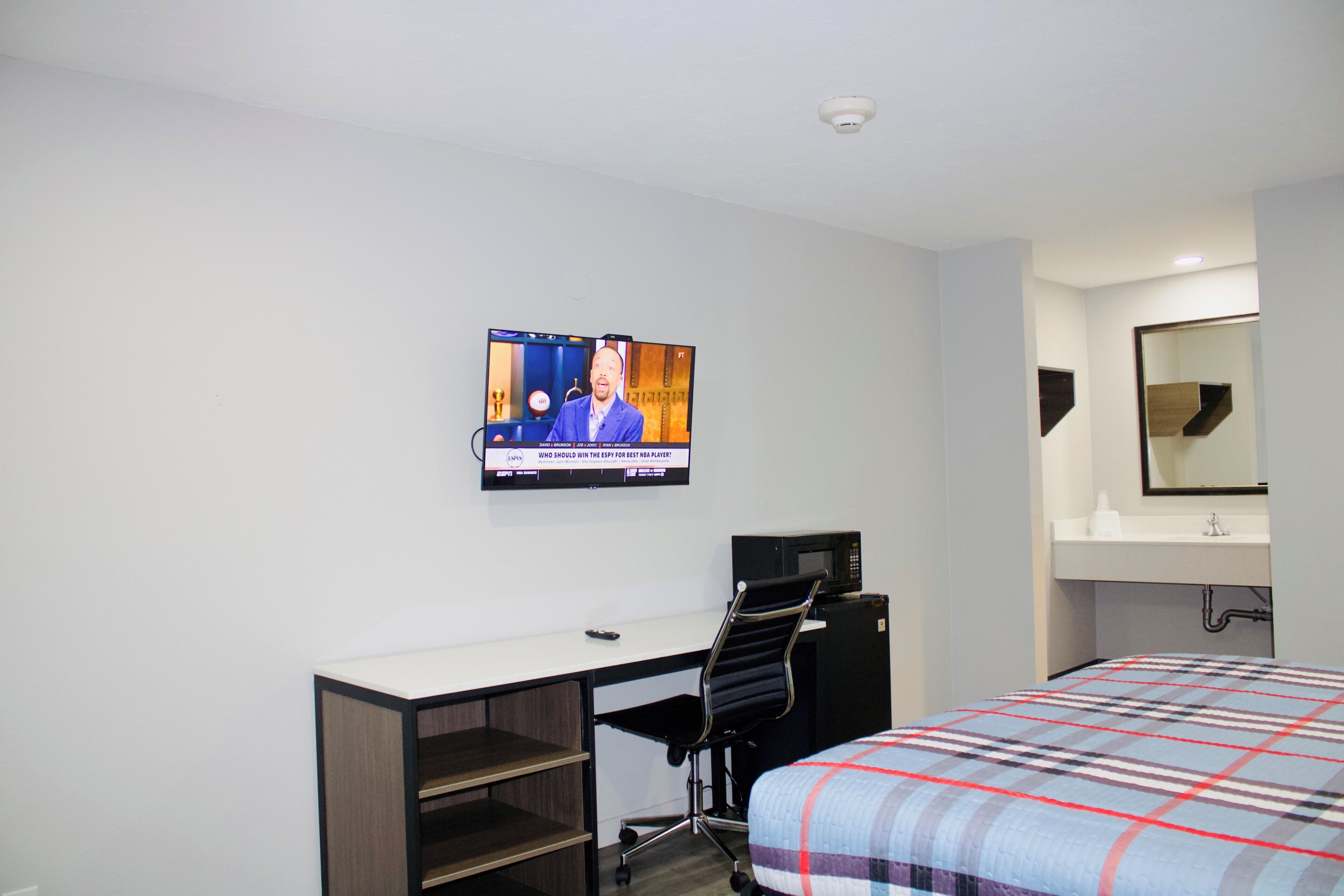 Una habitación de hotel con cama, escritorio con silla, televisor montado en la pared y baño con espejo y lavamanos.