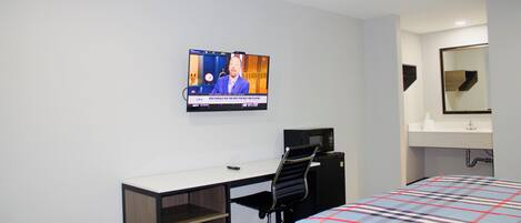 Una habitación de hotel con cama, escritorio con silla, televisor montado en la pared y baño con espejo y lavamanos.