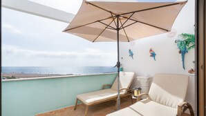 Un balcon avec vue sur la mer, un parasol beige, des chaises en osier et une table blanche.