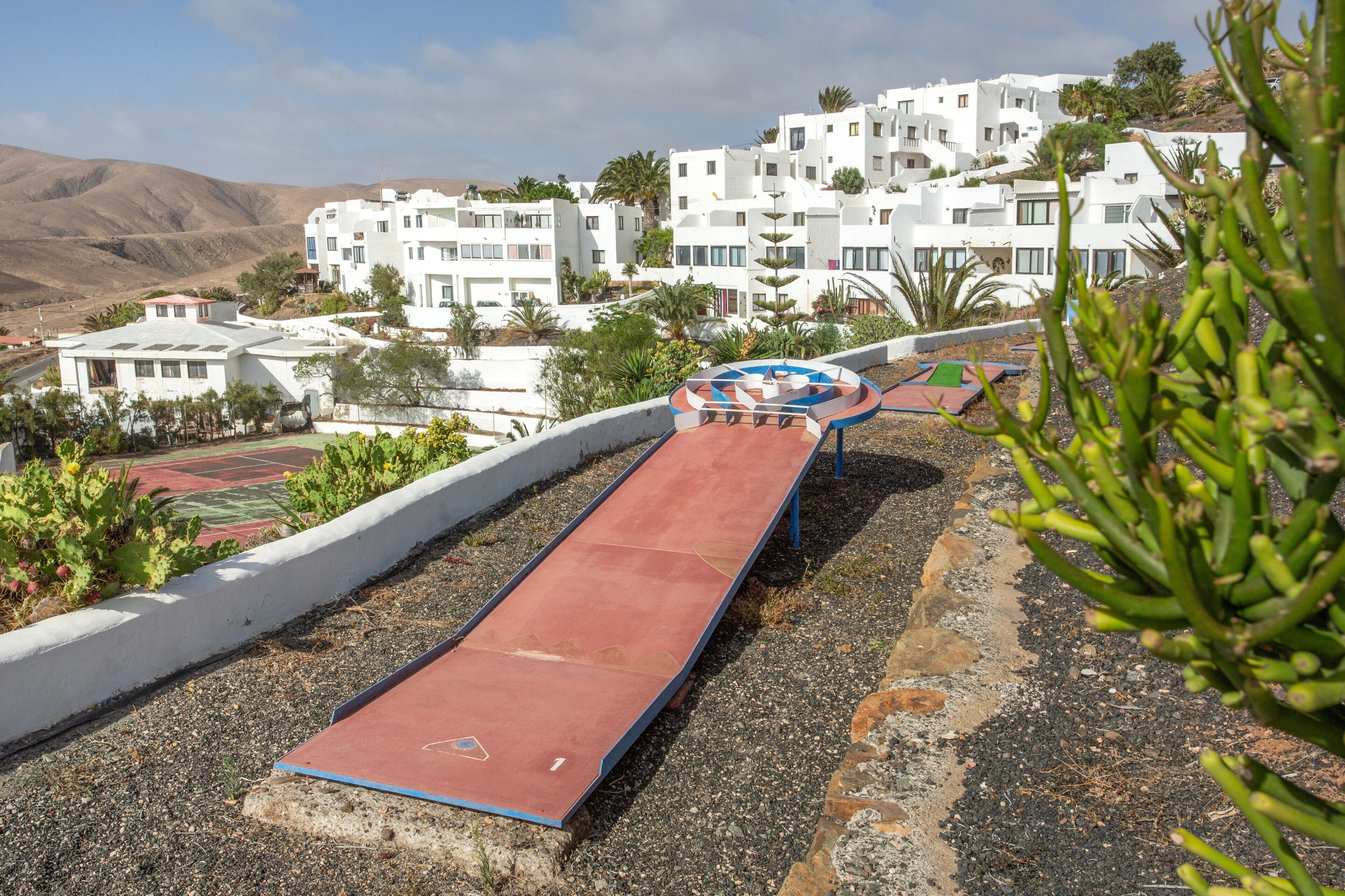 A red slide in a landscaped area with white buildings in the background.