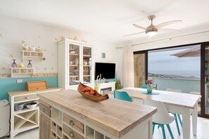 A modern kitchen with a wooden countertop, a dining area with a table and chairs, a TV, and a view of the sea.
