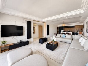 A modern living room with a flat-screen TV, a sofa set, a coffee table, and a kitchen area in the background.